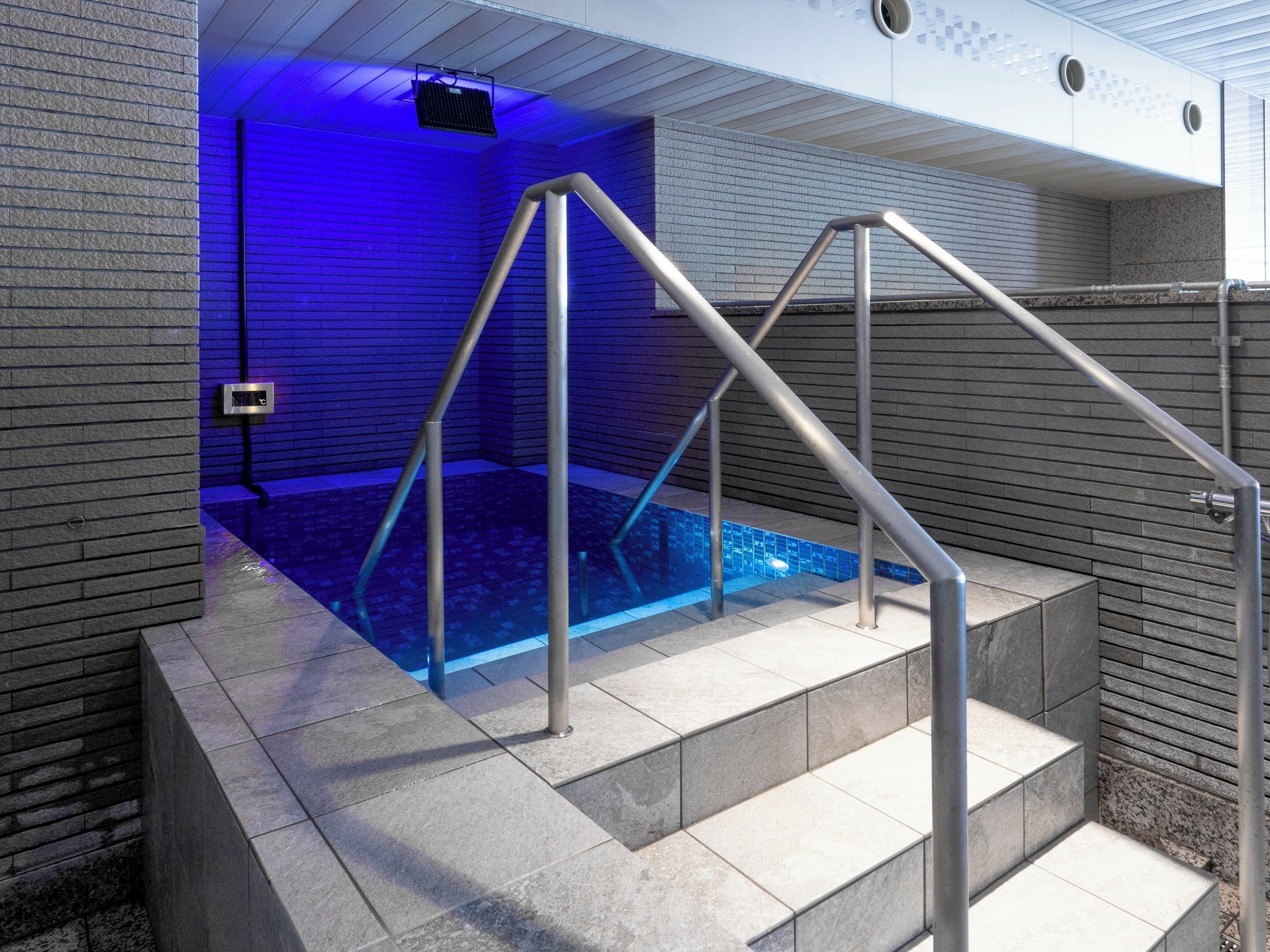 Un área de piscina interior moderna con una fuente iluminada en azul, barandales de metal y una pared de piedra.
