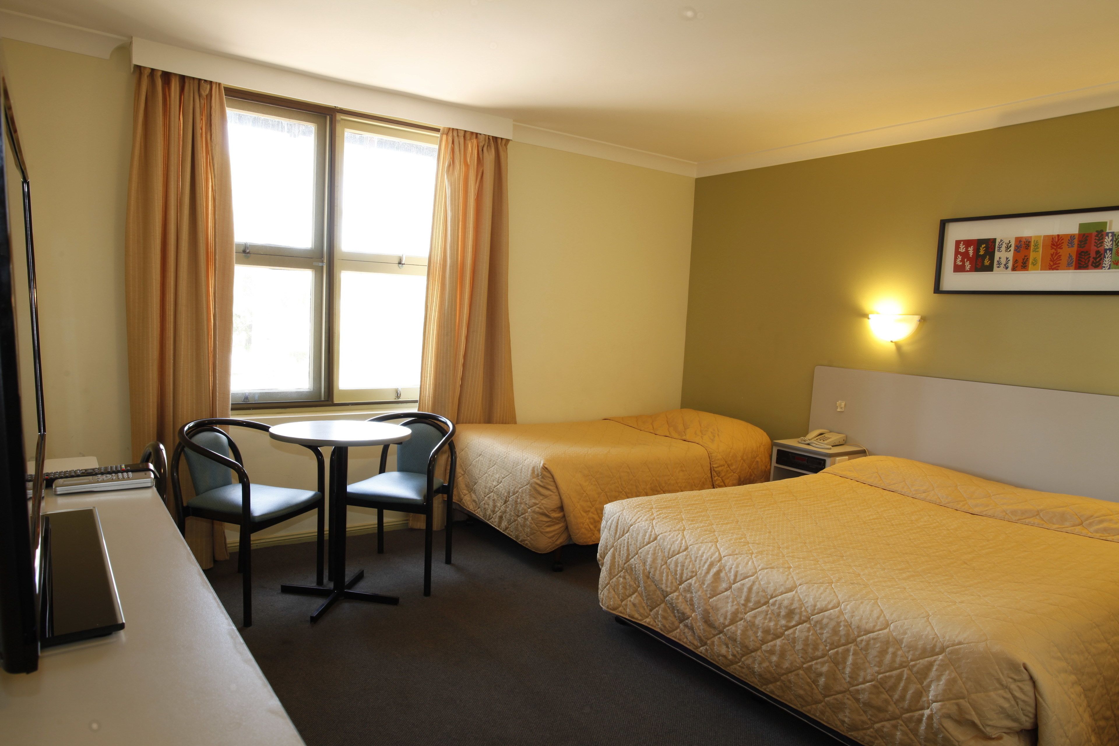 A hotel room with two beds, a desk with a chair, a small table, and a window with curtains.
