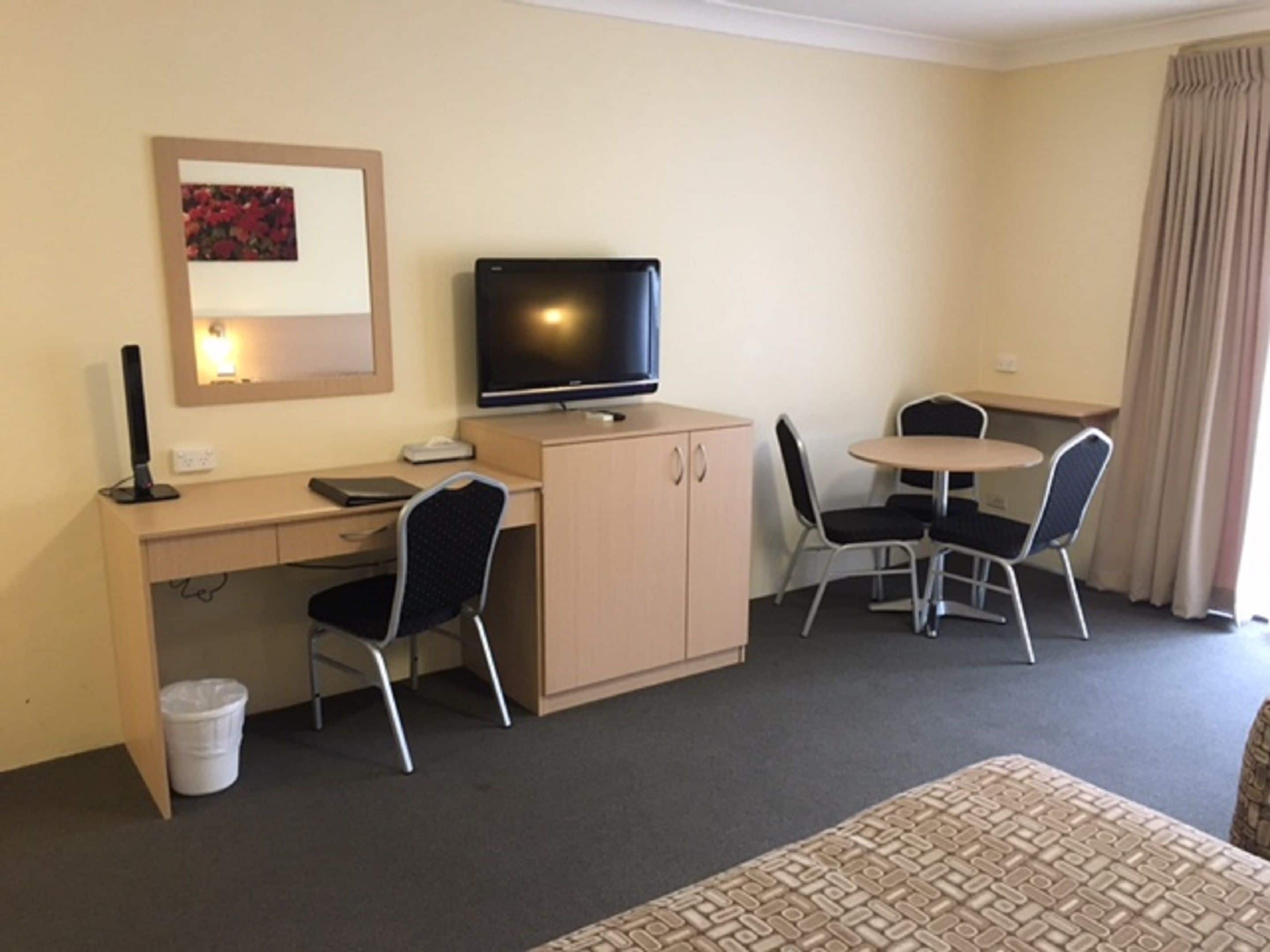 A hotel room with a desk, two chairs, a television, a small trash bin, and a bed.