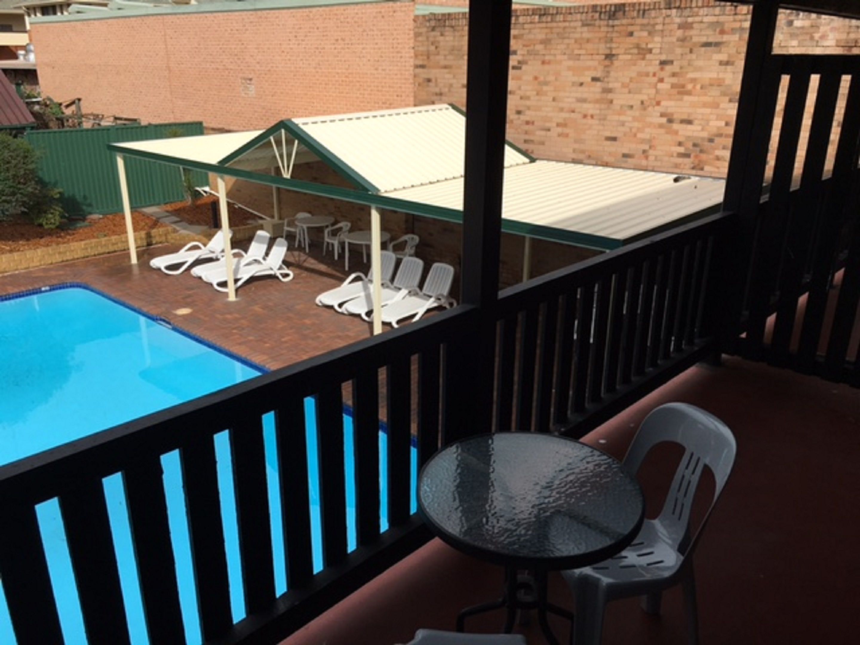 Zona de piscina con sillones de descanso y una mesa, rodeada por un muro de ladrillo y un cerramiento de vidrio.