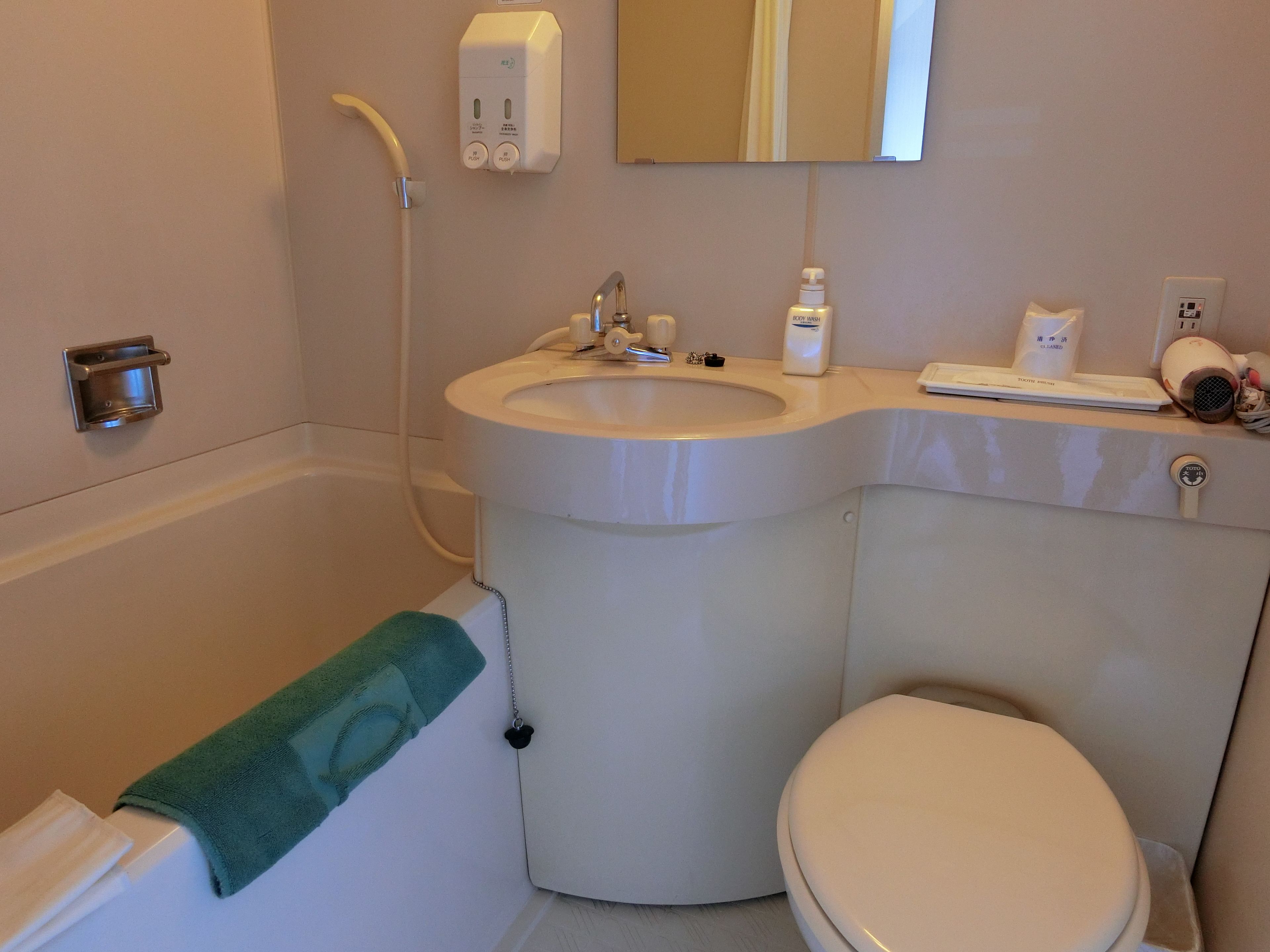 Un baño con bañera, lavabo e inodoro. Hay una toalla colgada de la bañera, un dispensador de jabón y un secador de pelo sobre el mostrador del lavabo.