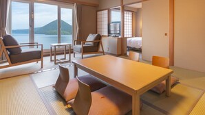 Hotelkamer met een groot raam met uitzicht op een meer en bergen, een eethoek met tatami matten, en een televisie in de hoek.