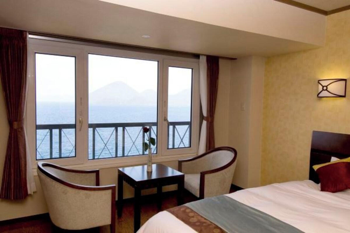 Ein Hotelzimmer mit einem Bett, zwei Stühlen, einem kleinen Tisch und einem großen Fenster mit Blick auf das Meer und die Berge.