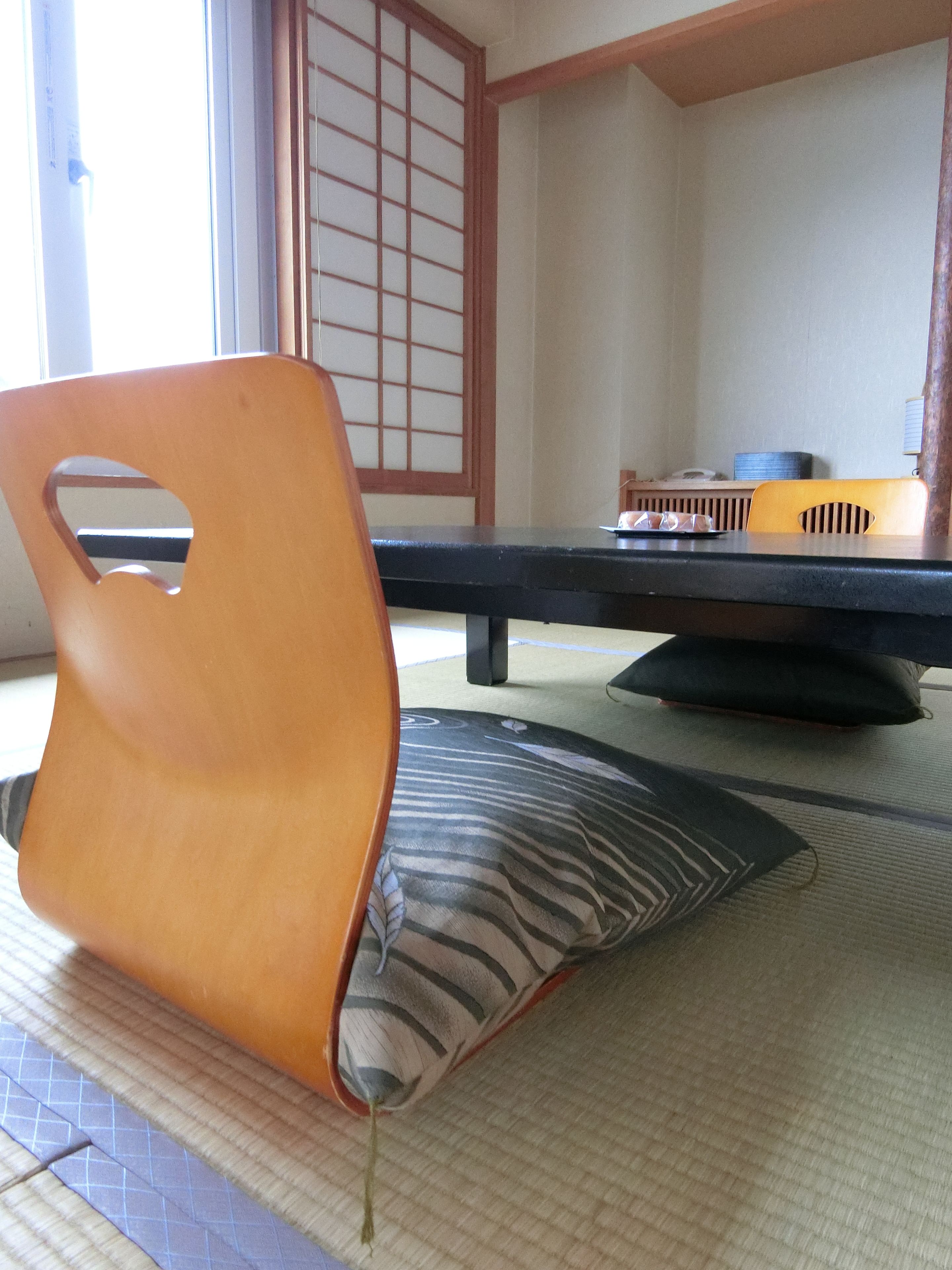 Una habitación con esteras de tatami, una mesa baja, una silla de madera y un cojín.