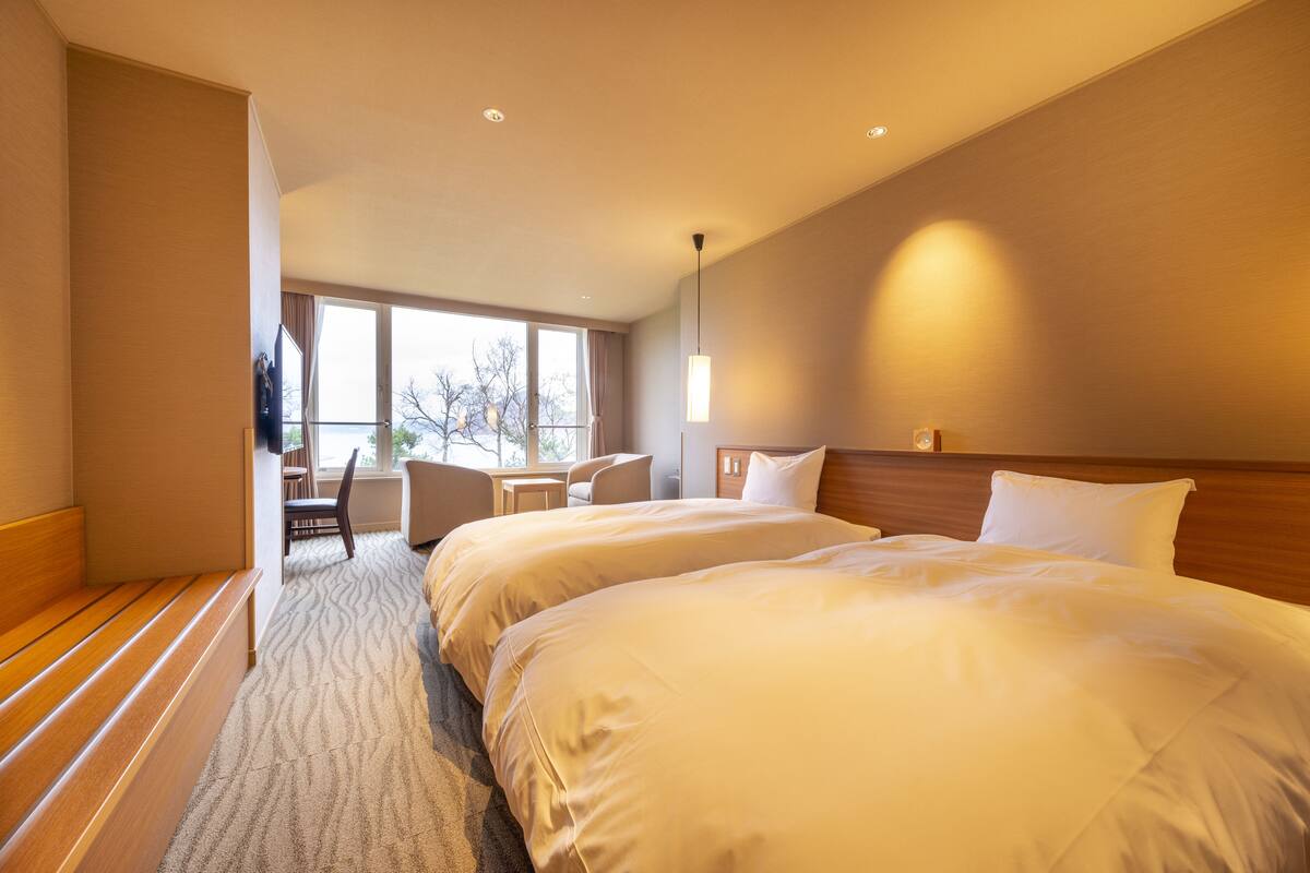 Une chambre d’hôtel avec deux lits, un banc en bois, un bureau avec une chaise et une grande fenêtre donnant sur la forêt.