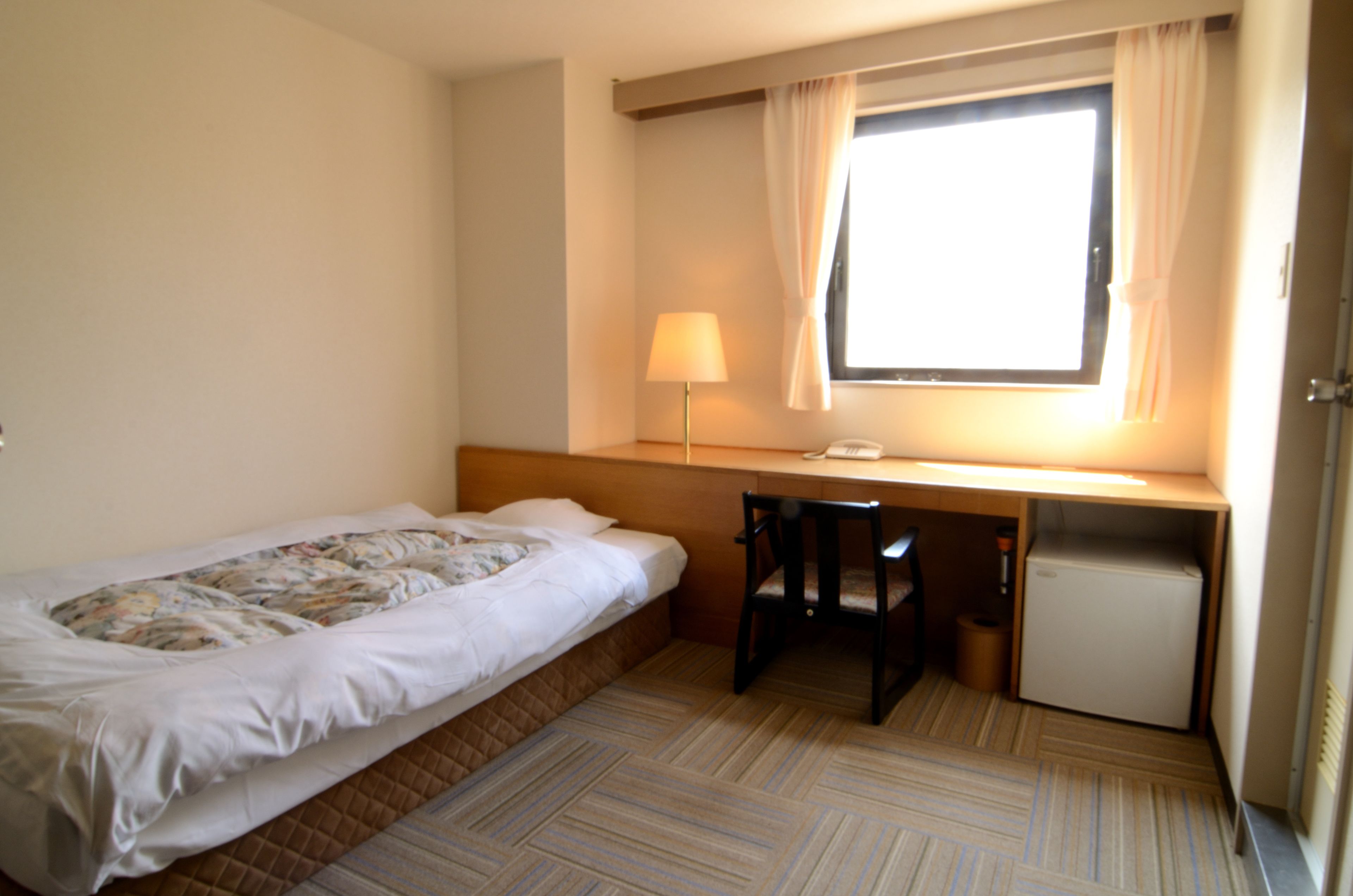 Una habitación de hotel pequeña con una cama individual, un escritorio, una silla, una lámpara y un refrigerador pequeño.