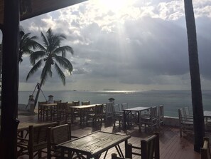 A seaside restaurant with outdoor seating, palm trees, and a view of the ocean.