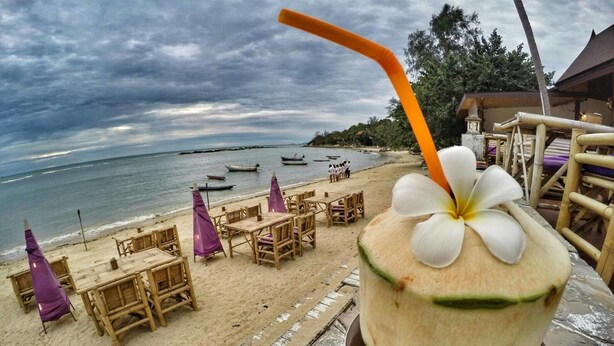 A beachside restaurant with wooden tables and chairs, a coconut drink with a straw, and a view of the ocean and boats.