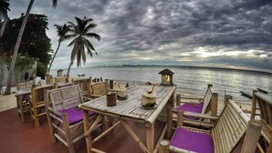 A wooden deck with bamboo furniture and purple cushions, overlooking a calm sea with palm trees and a cloudy sky.
