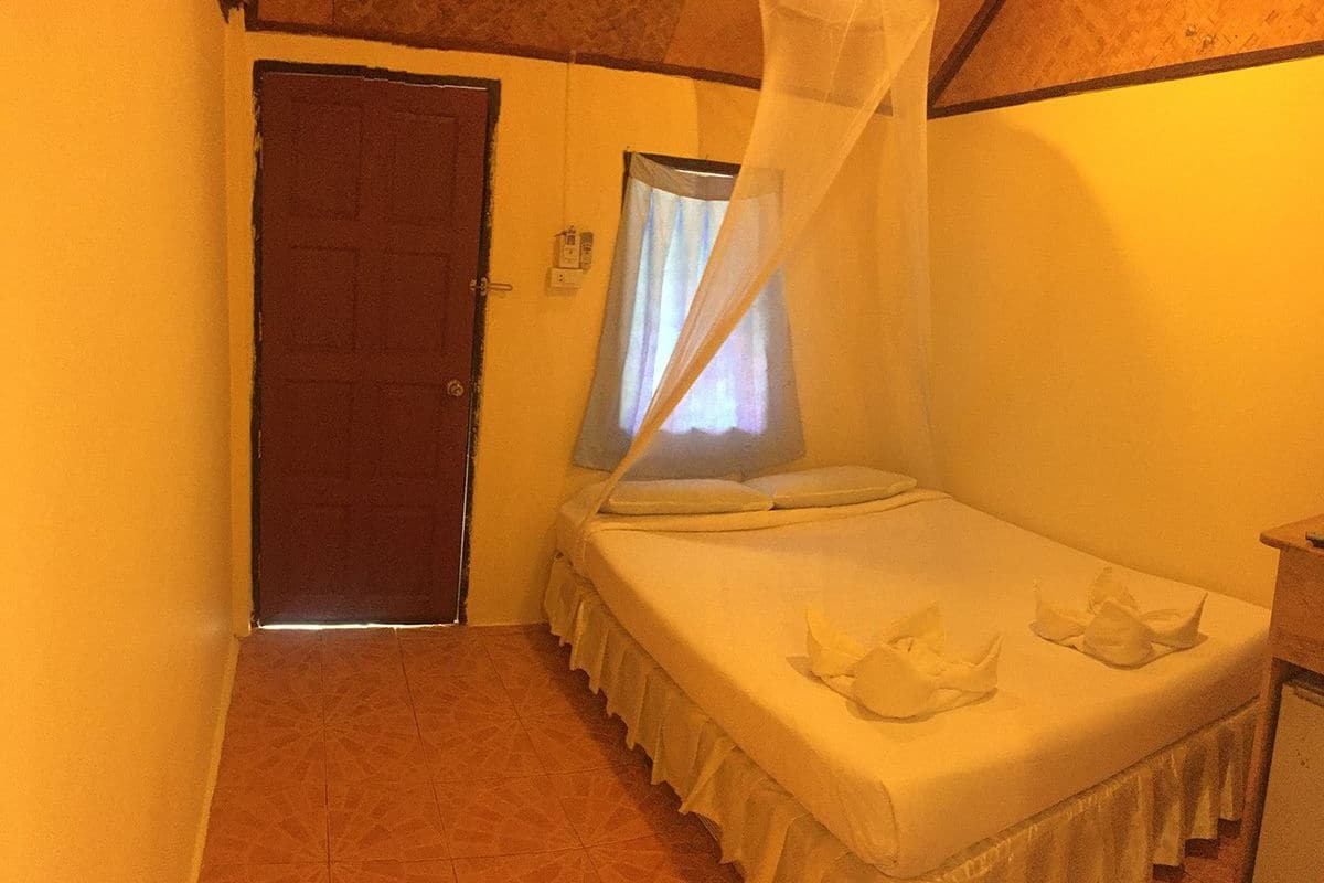 Ein kleines Zimmer mit einem Einzelbett, einem Moskitonetz darüber, einer Tür und einem Fenster mit Vorhängen.