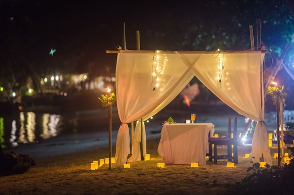 A tented outdoor dining area with a table and chairs, surrounded by candles and string lights.