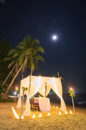 A beachside tent with a table and chairs, surrounded by candles and palm trees, under a night sky with a visible moon.
