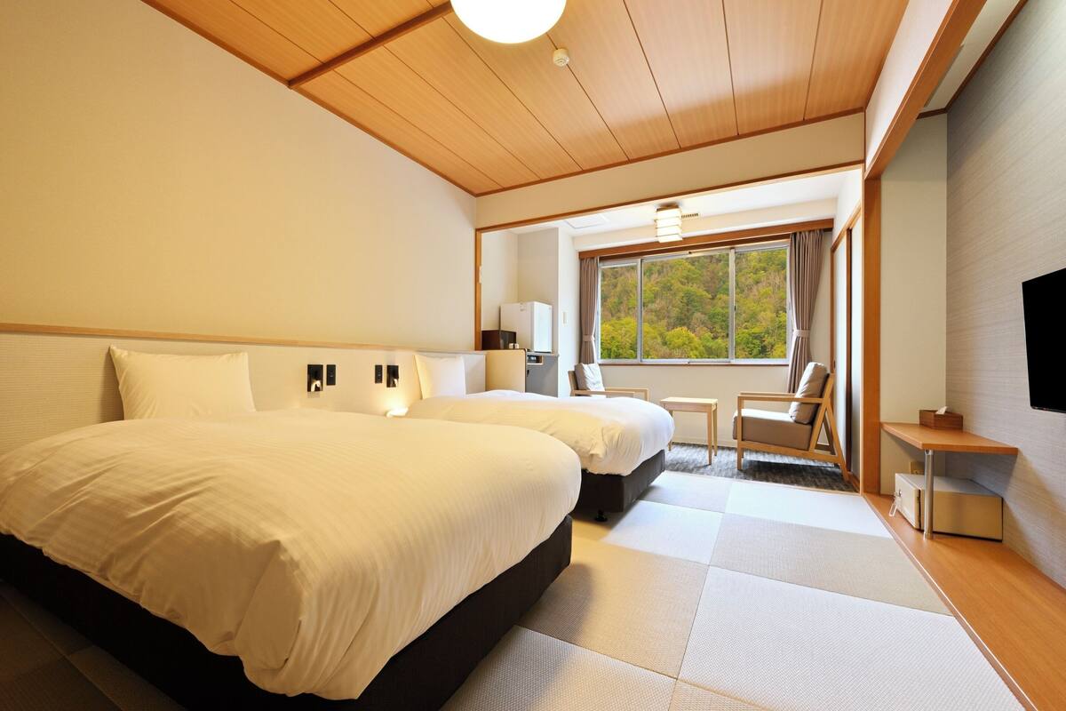 A hotel room with two beds, a small table, a chair, a TV, and a window with a view of trees.