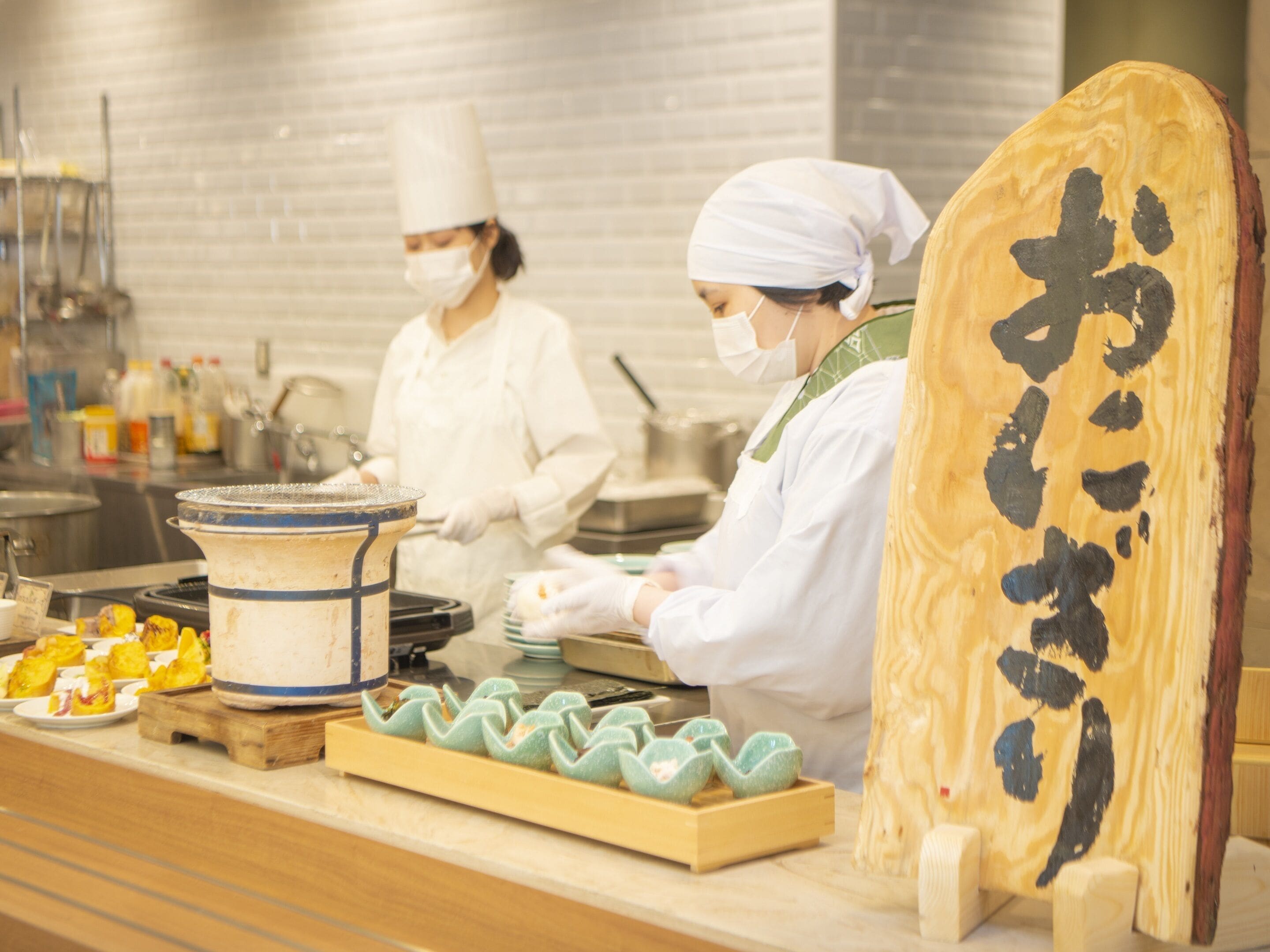 A kitchen with chefs preparing food, a large wooden board with calligraphy, and a tray of food items.