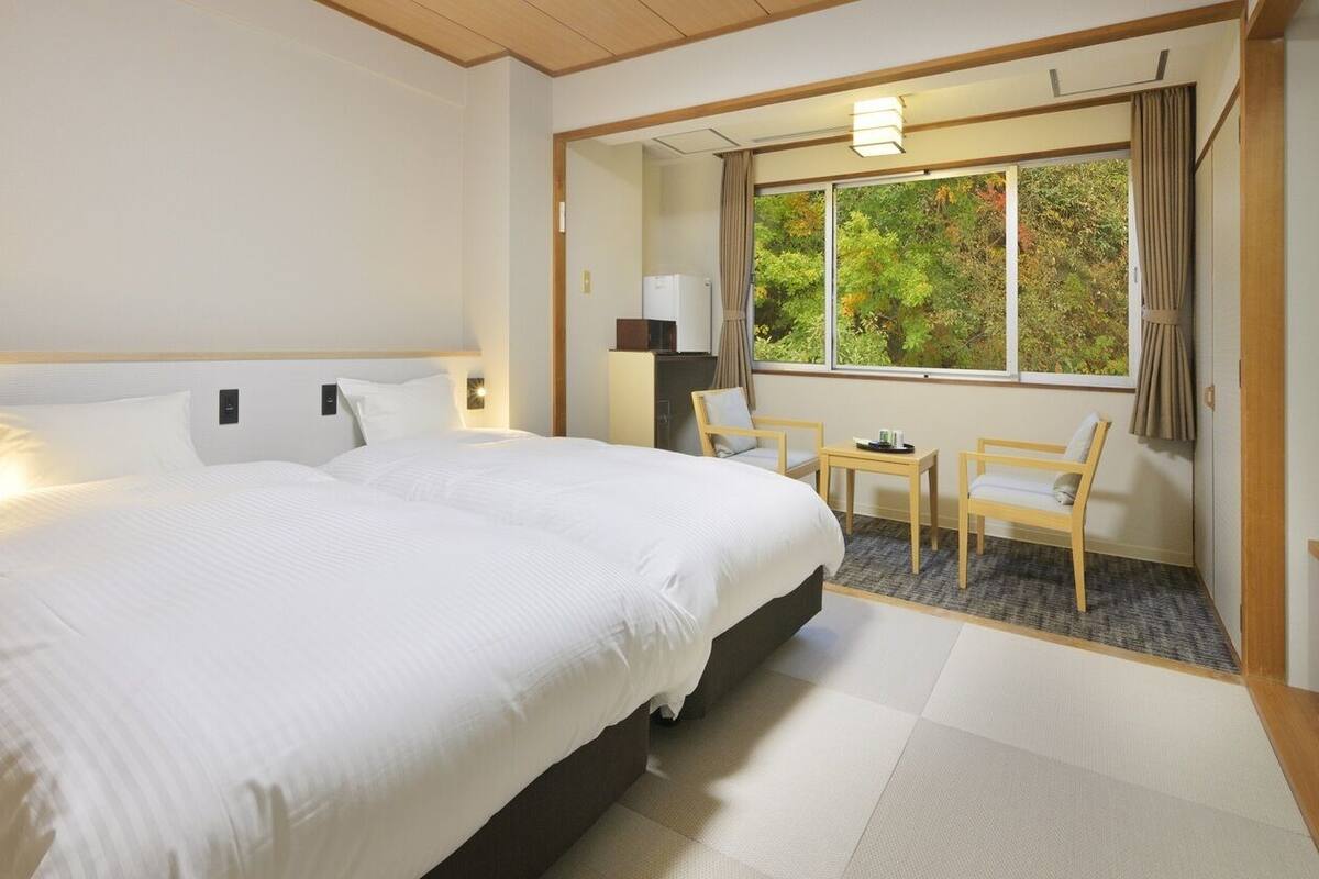 A hotel room with a large bed, a small table, two chairs, a window with a view of trees, and a wooden ceiling.