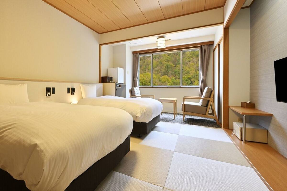 A hotel room with two beds, a small table, a chair, a window with a view of trees, and a TV mounted on the wall.