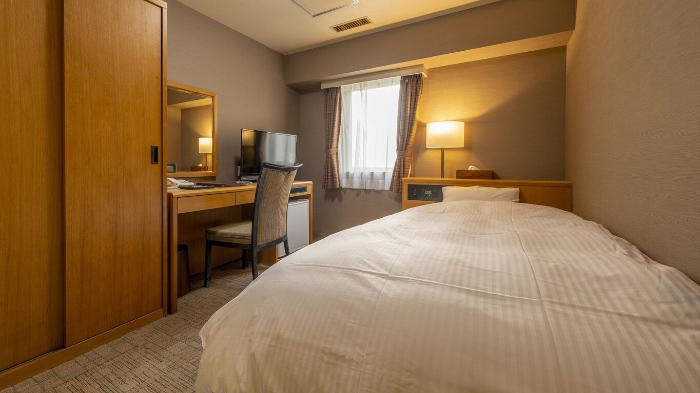 Habitació d'hotel amb un llit, una taula de treball amb una cadira, una televisió i una finestra amb cortines.