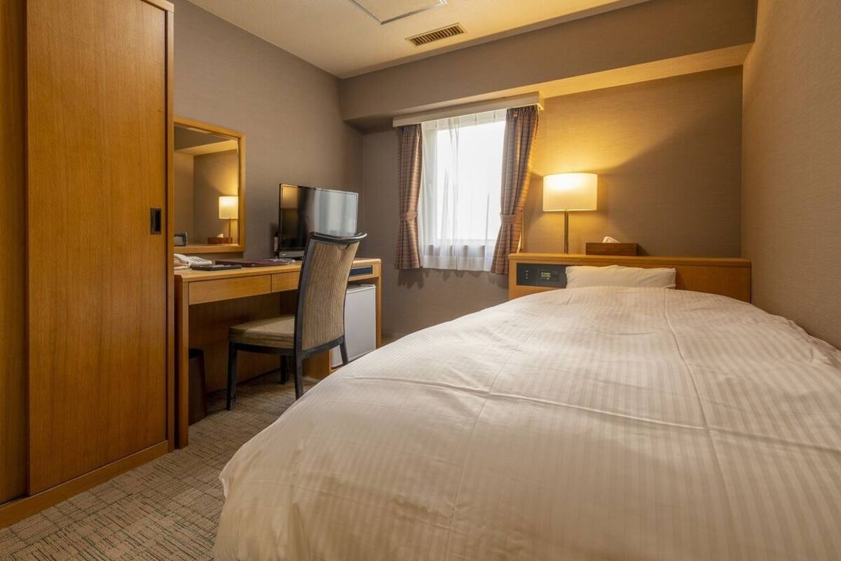 A hotel room with a bed, a desk with a chair, a television, and a window with curtains.