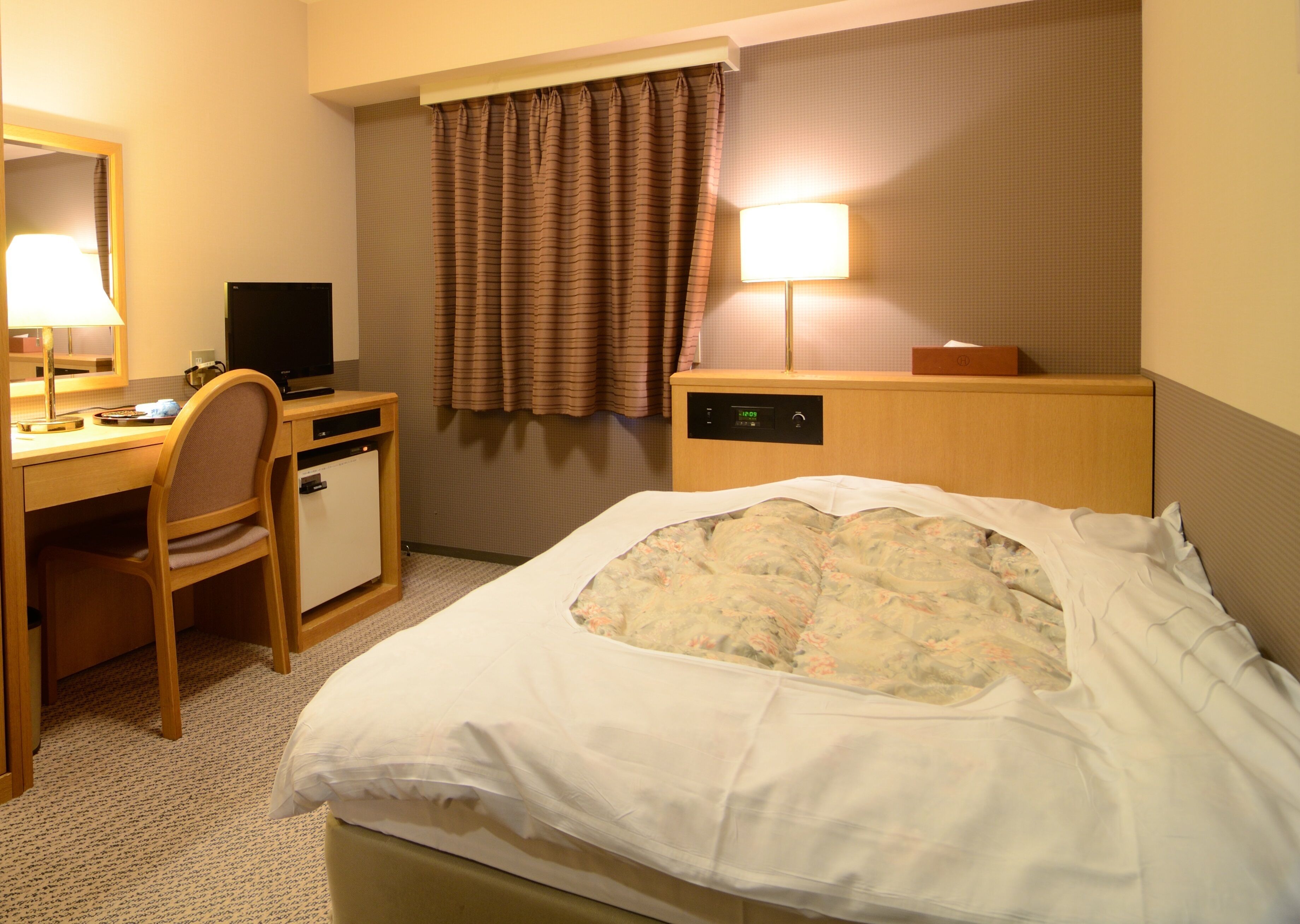 A hotel room with a bed, a desk, a chair, a lamp, a nightstand, and a window with curtains.