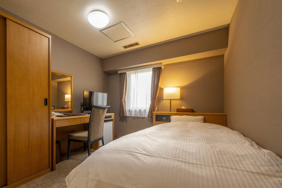 A hotel room with a bed, desk, chair, television, and a nightstand with a lamp.