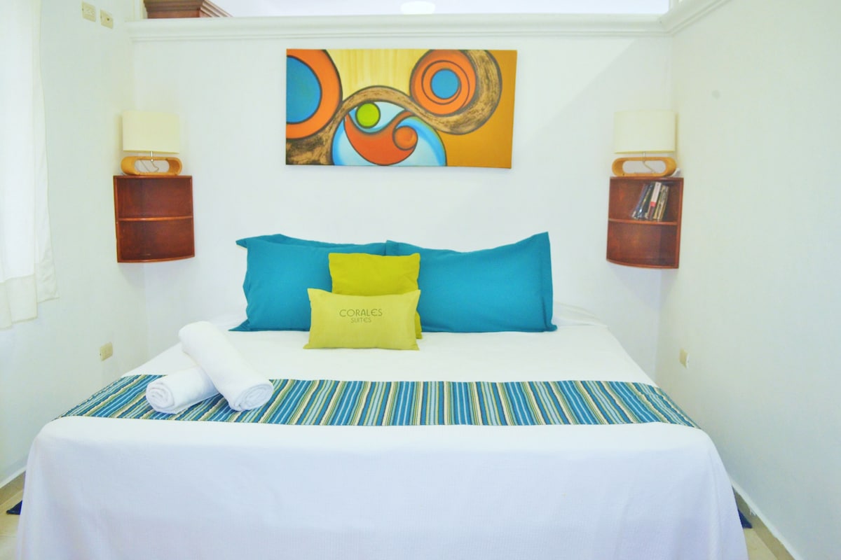 A hotel room with a bed, pillows, a striped bedspread, a painting on the wall, and two bedside tables with lamps.