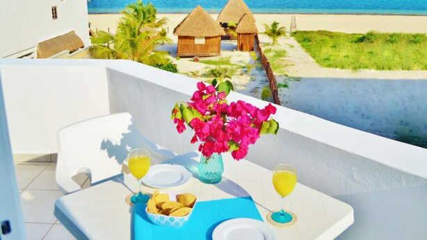 A balcony with a table set for two, overlooking a beach with thatched-roof huts and a clear blue sea. - Corales Suites (Puerto Morelos)