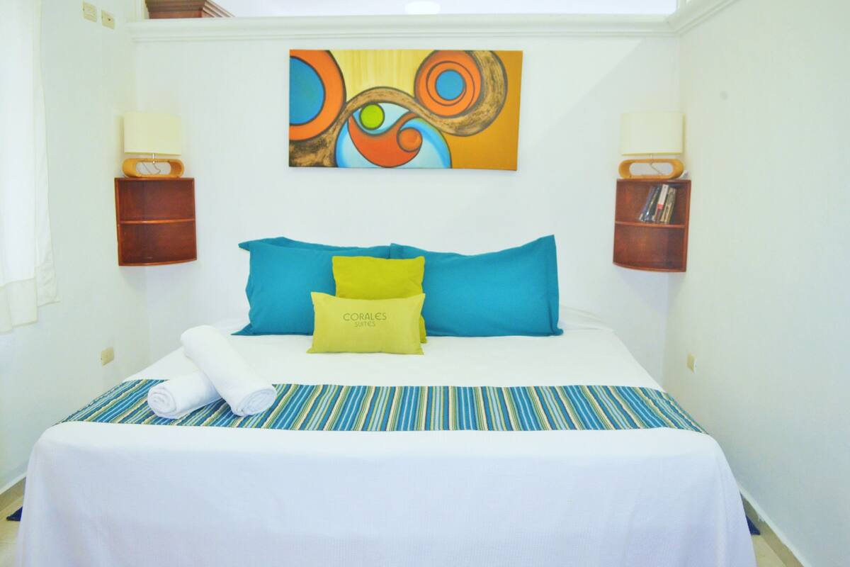 Habitación de hotel con cama, almohadas, cubrecama a rayas, un cuadro en la pared y dos mesitas de noche con lámparas.