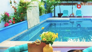 A poolside area with a blue-tiled pool, a wooden table with a yellow vase and a brown bag, and two blue lounge chairs. - Corales Suites (Puerto Morelos)