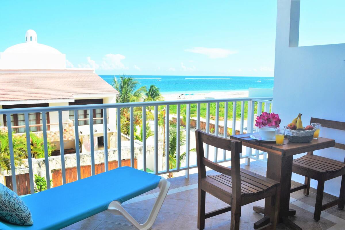 Un balcón con vista a la playa, una mesa con fruta y flores, y una silla azul.