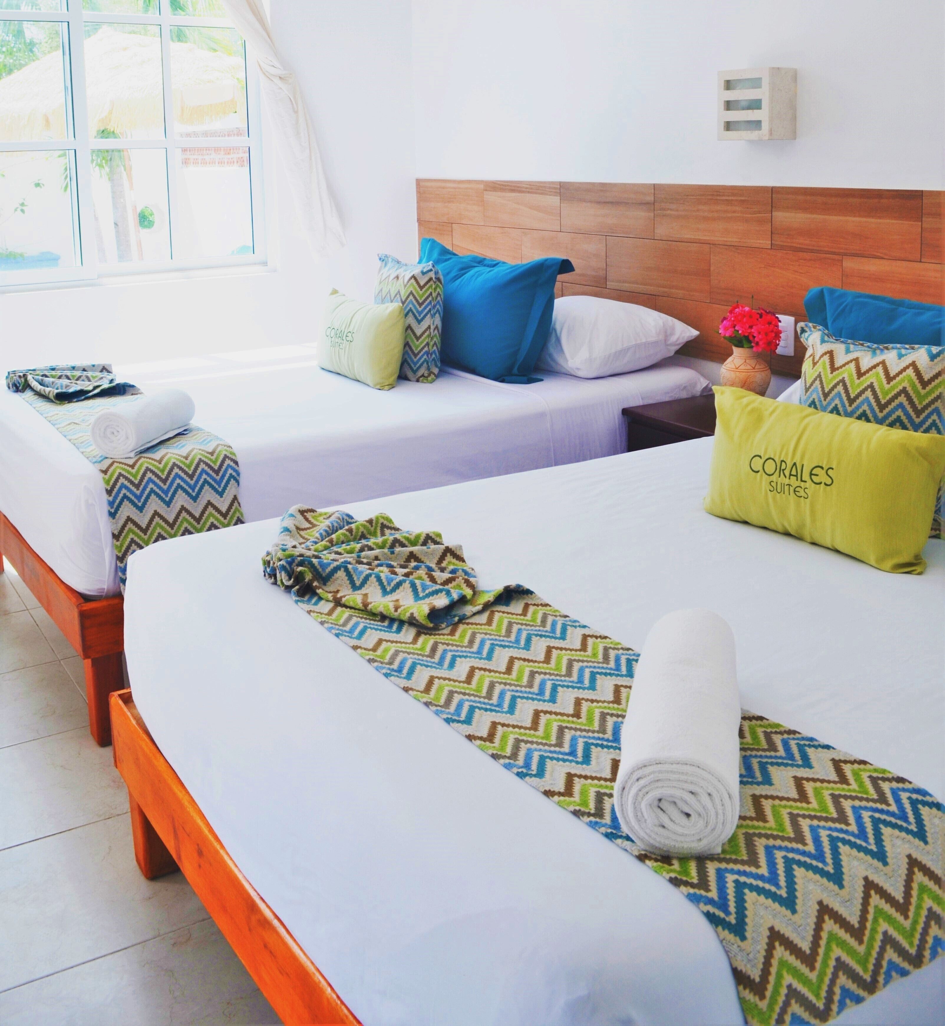 A hotel room with two beds, each with a wooden headboard, white linens, and colorful patterned blankets. There are pillows with text and a vase with flowers on the nightstand.