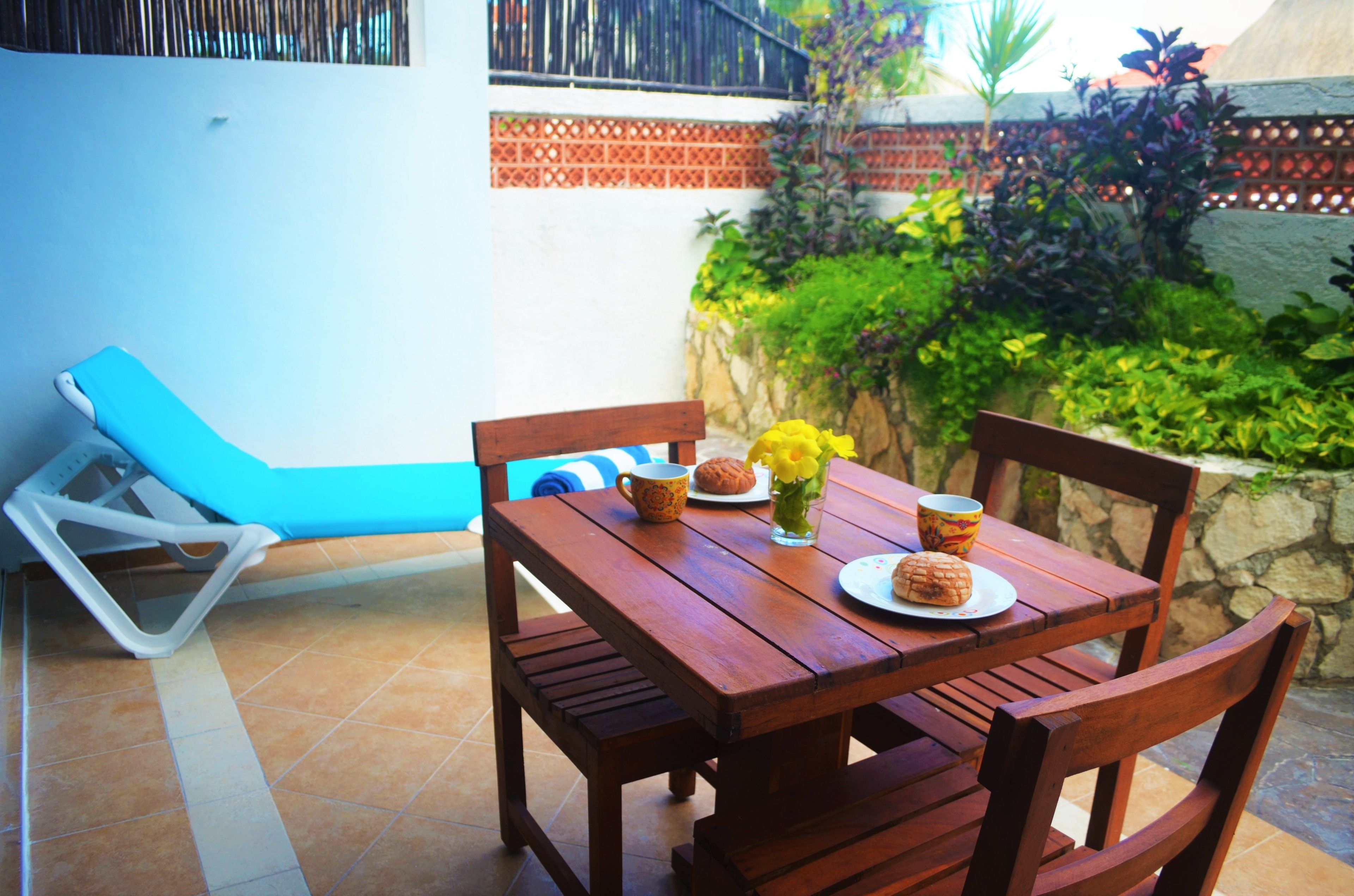 A wooden table with chairs, a blue lounge chair, and a small garden area with plants and a stone wall.