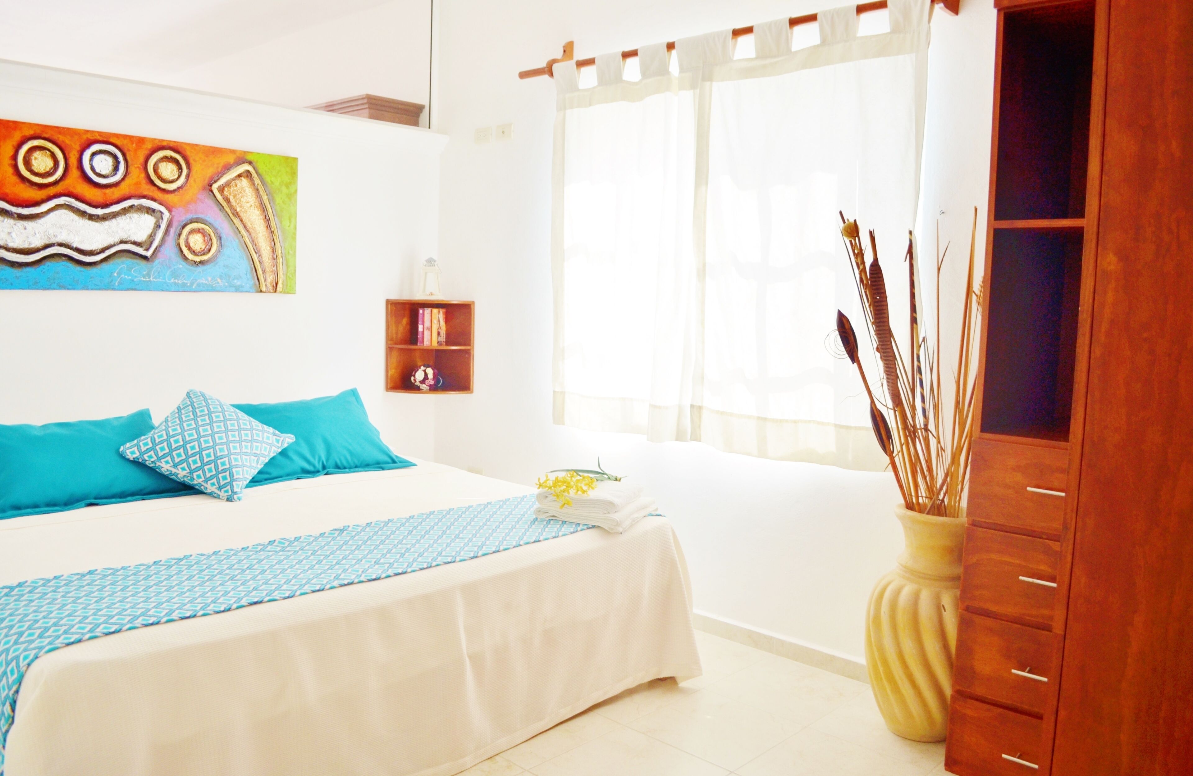 Quarto de hotel com cama, mesa de cabeceira, um vaso com plantas e um quadro na parede.