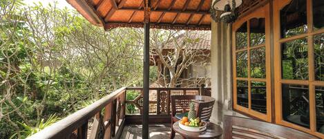 A wooden balcony with a table holding fruits and a chair, overlooking a lush garden.