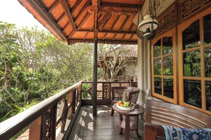 Un balcón de madera con una mesa que sostiene frutas y una silla, con vistas a un jardín exuberante.