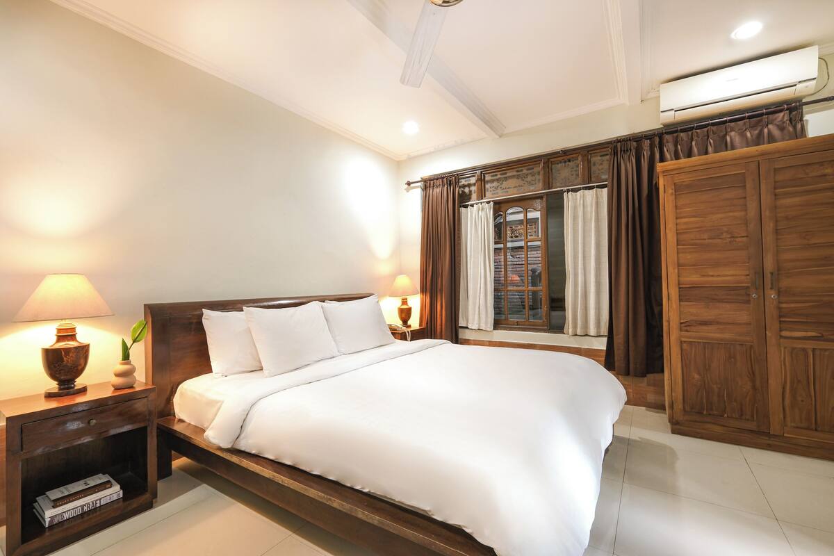 A neatly made bed with white linens, a wooden nightstand with a lamp, and a wardrobe with a mirror.