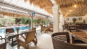 Zona de piscina interior con mobiliario de madera y techo de paja.