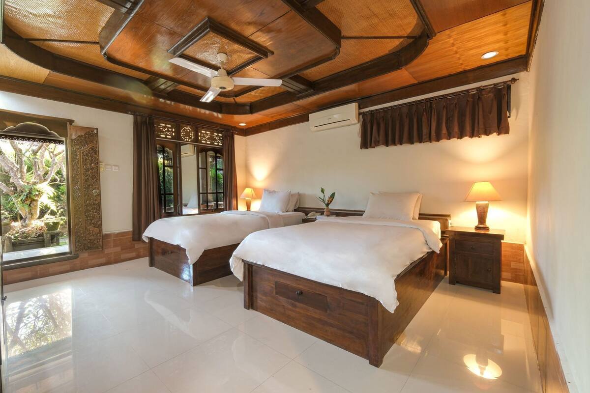 A bedroom with two wooden beds, a ceiling fan, an air conditioning unit, and a view of greenery outside.