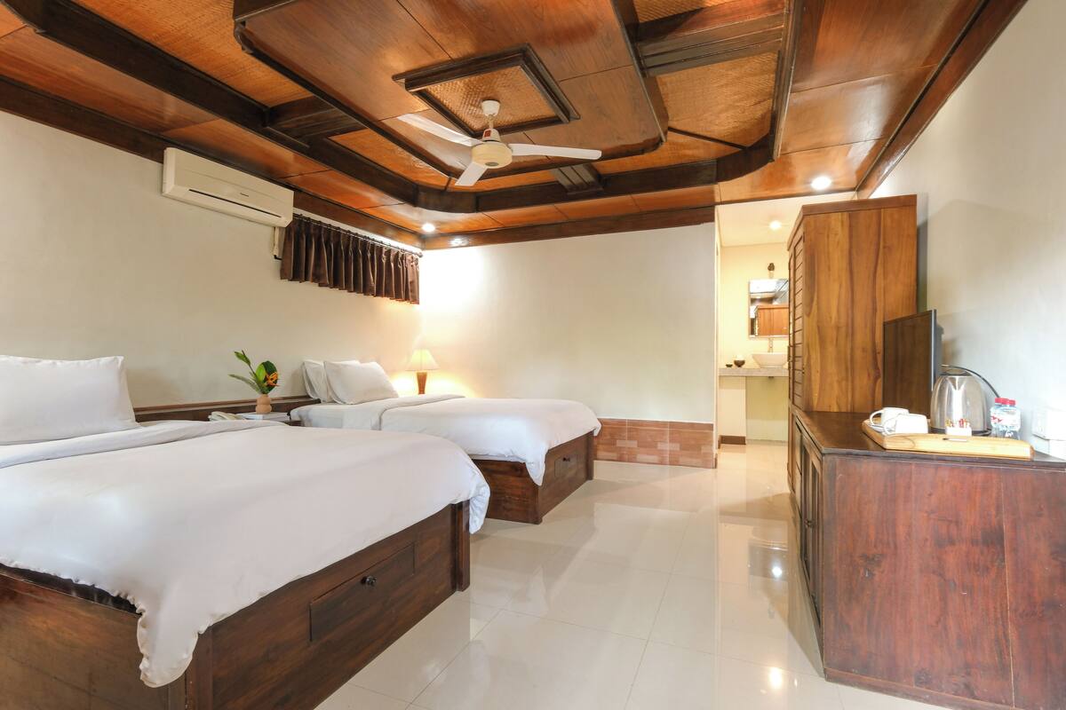 A hotel room with two beds, a wooden ceiling, an air conditioning unit, a desk with a mirror, and a bathroom visible through an open door.