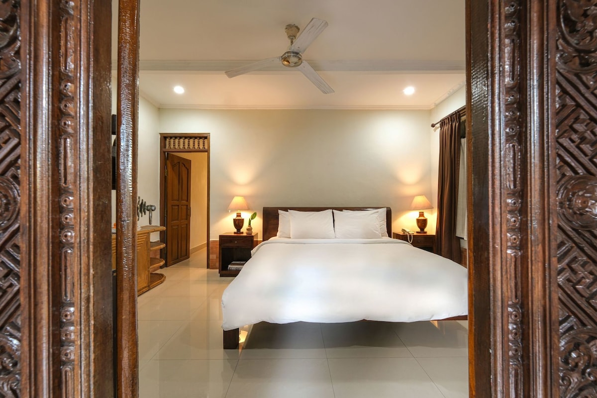 A hotel room with a large bed, two bedside tables with lamps, a ceiling fan, and a wooden door frame.
