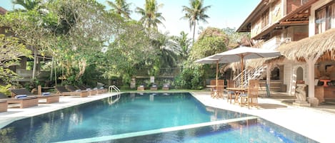 A swimming pool with lounge chairs and umbrellas, surrounded by tropical vegetation and a building with a thatched roof.