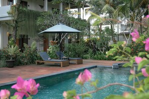 A poolside area with lounge chairs and an umbrella, surrounded by lush greenery and a clear pool.