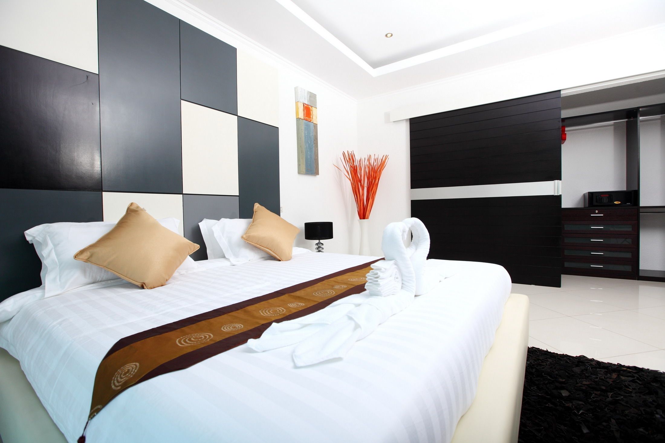 Quarto de hotel moderno com uma cama grande, uma mesinha de cabeceira, um criado-mudo, um painel de parede preto e branco, um quadro e um banheiro visível através de uma porta aberta.