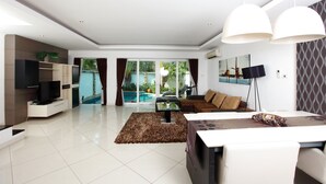 A modern living room with a TV, sofa, dining table, and large windows overlooking a pool. - Hidden Palm Pool Villas (Pattaya)