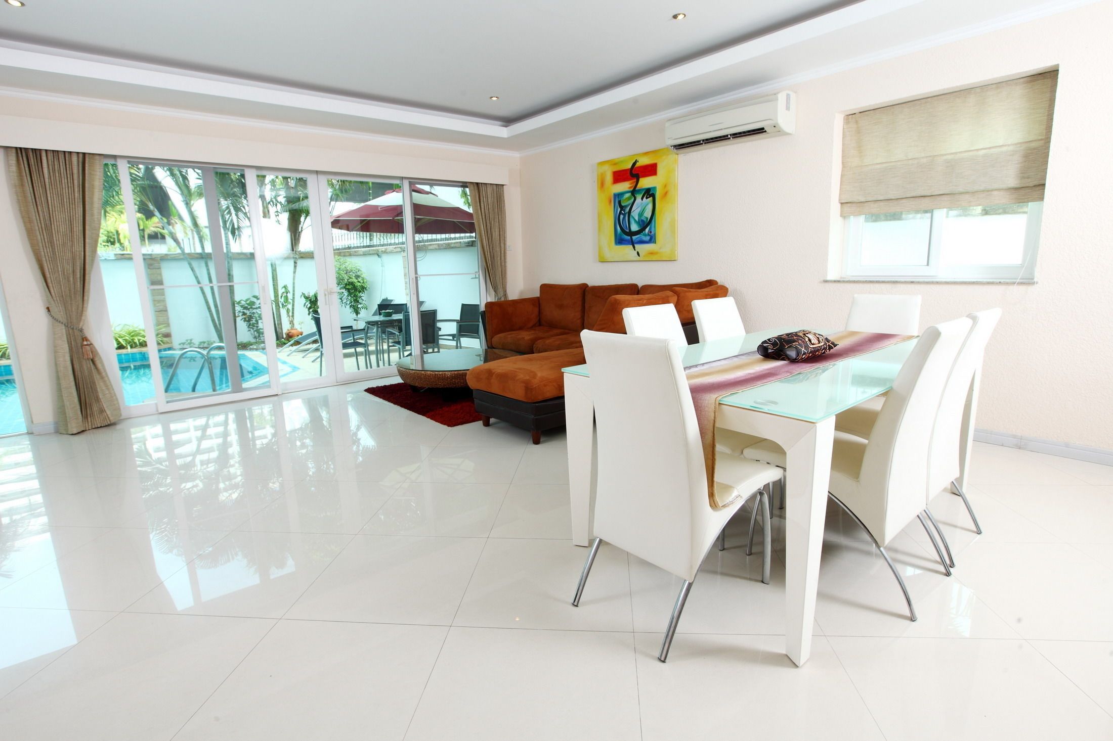 A spacious living room with a glass dining table and white chairs, a sofa, and a large window overlooking a pool area.