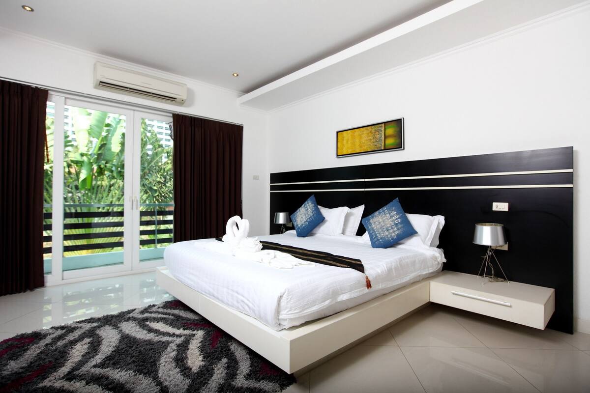 A modern bedroom with a large bed, a nightstand, a lamp, and a view of greenery through the window.