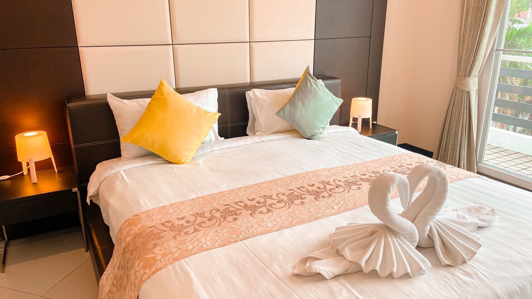 Een net opgemaakt bed met een decoratieve zwaan van handdoek, twee nachtkastjes met lampen, en een raam met gordijnen.