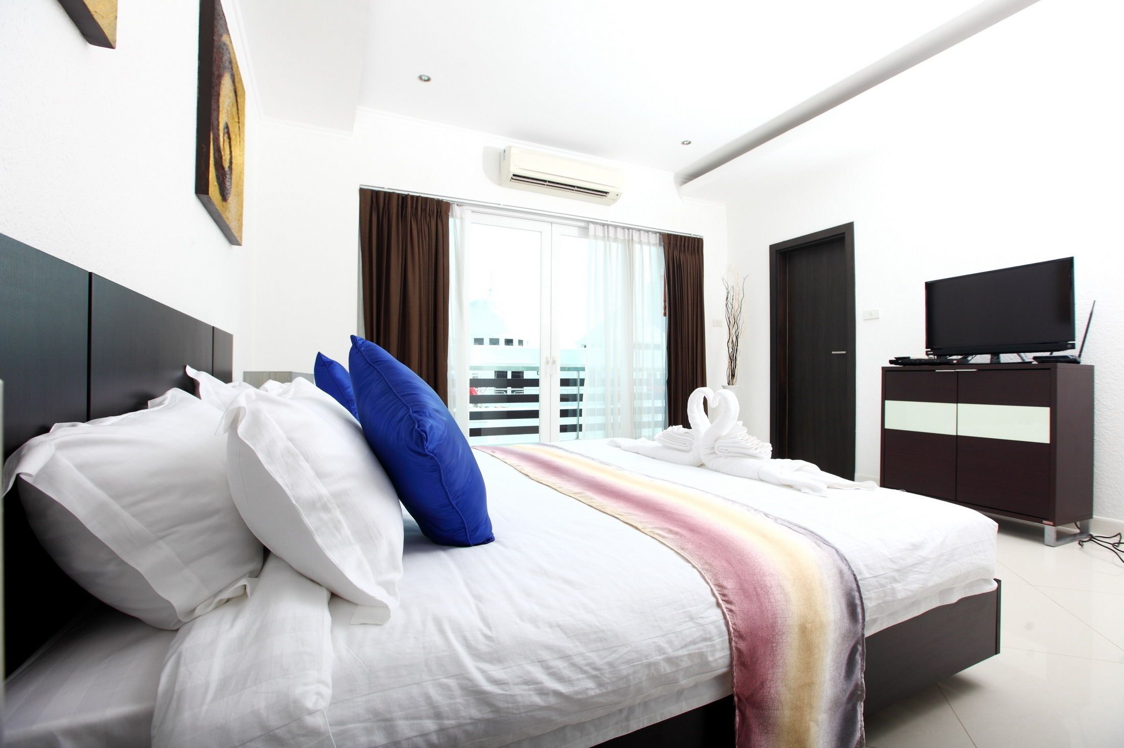 A modern hotel room with a large bed, a flat-screen TV, a wardrobe, and a view of the city through the window.