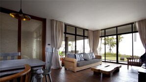 A spacious living area with a wooden floor, a sofa with cushions, a dining table with chairs, and large windows offering a view of the outdoors. - Na Tara Resort (Ko Chang)