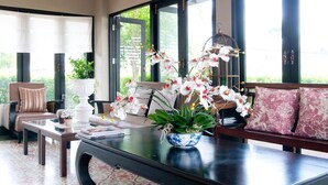 A well-lit room with a sofa, a coffee table, and a vase with flowers. There are windows with curtains and a view of greenery outside. - Ploen Pattaya Residence by Tolani (Pattaya)