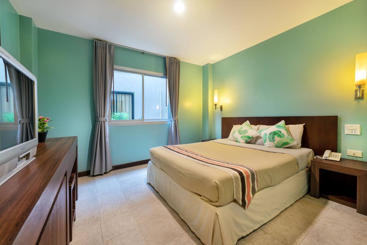A hotel room with a bed, a TV, a bedside table with a phone, a window with curtains, and a wall-mounted light.